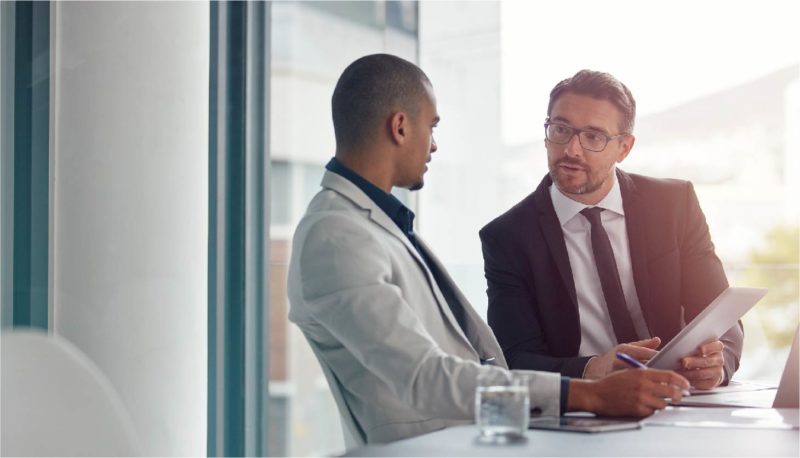 Two businessmen sit at an office desk and talk with each other
