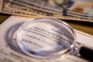 Severance Pay agreement document on wooden board with a magnifying glass on top of it and a stack of US currency above it.