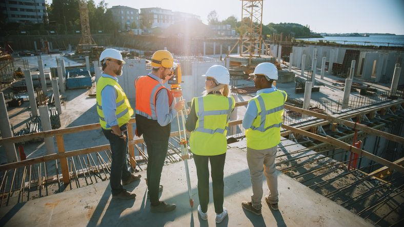 Construction Worker Using Theodolite Surveying Optical Instrument for Measuring Angles in Horizontal and Vertical Planes on Construction Site. Engineers and Architect Discuss Plans Next to Surveyor.
