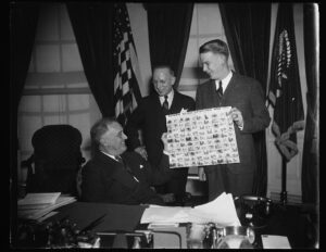 Two man are standing next to President Franklin Roosevelt, who is seated at his desk, and one man is holding up a sheet of stamps that says "National Wildlife Restoration Week" at the top.