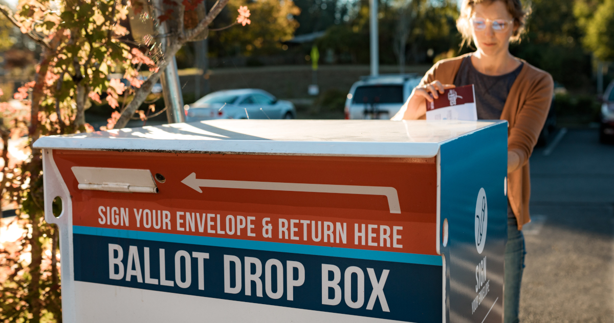 A Woman Places A Ballot In A Ballot Drop Box