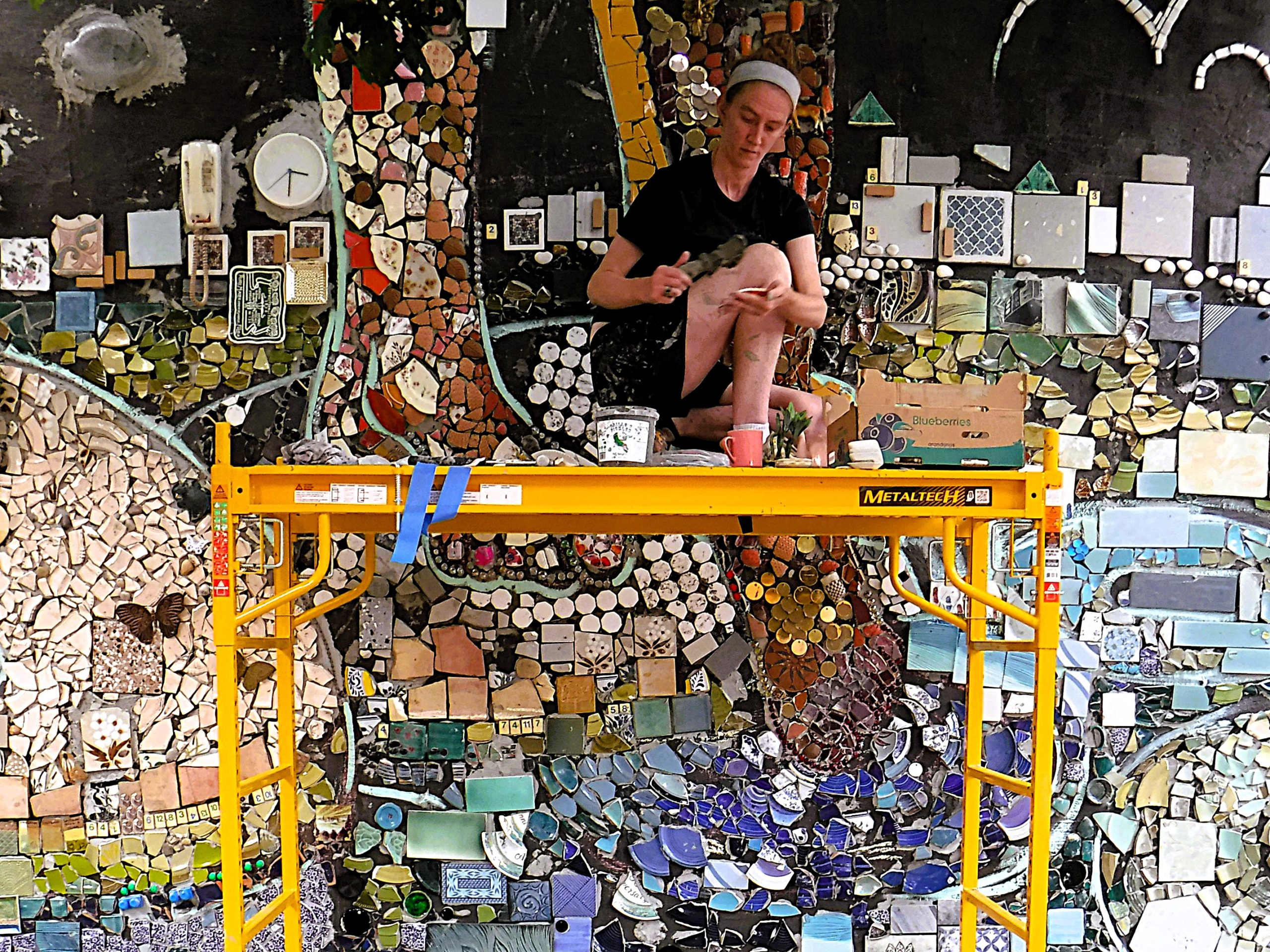 A person sits on scaffolding while working on a colorful mosaic mural, with various tiles and materials surrounding them.
