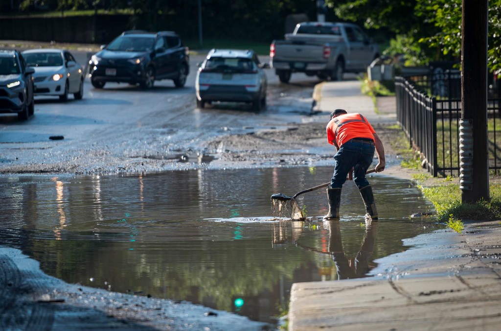 A worker in an orange vest stands in a large puddle on the side of a road, scooping debris with a net. Cars are passing by on the wet, muddy street.