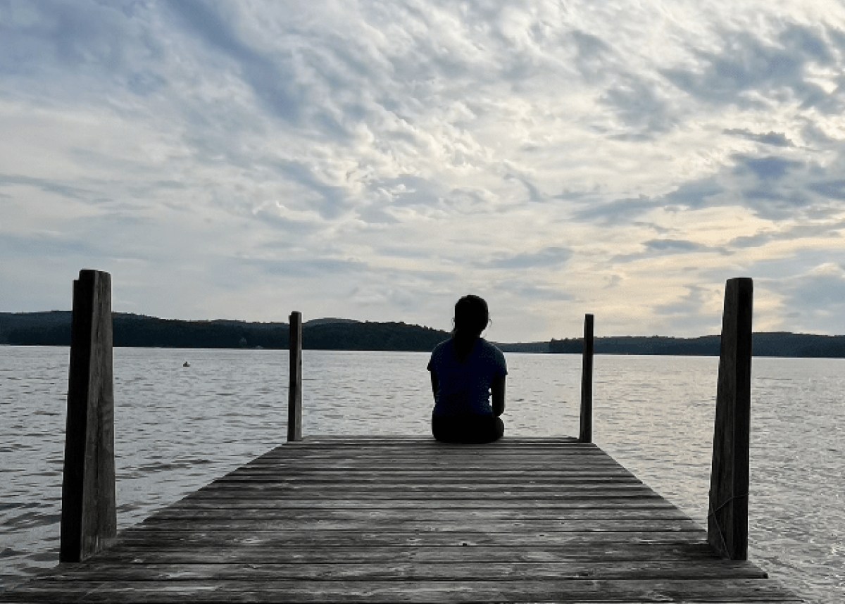 A person sits at the end of a wooden dock, facing a tranquil lake under a cloudy sky.