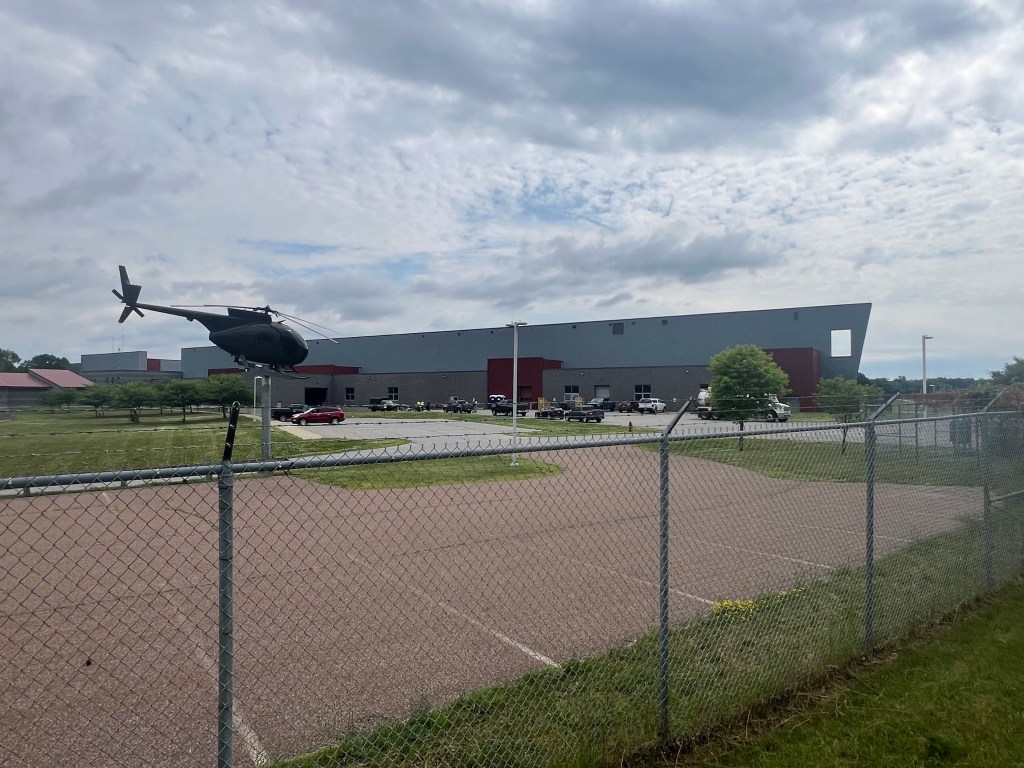 A helicopter hovers near a chain-link fence with a large industrial building and parked vehicles in the background under a partly cloudy sky.