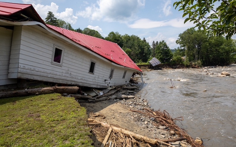 A small building with a red roof lies partially collapsed in a large stream of water. This is a motel in Hardwick, Vermont that suffered from the devastating flooding in 2023.