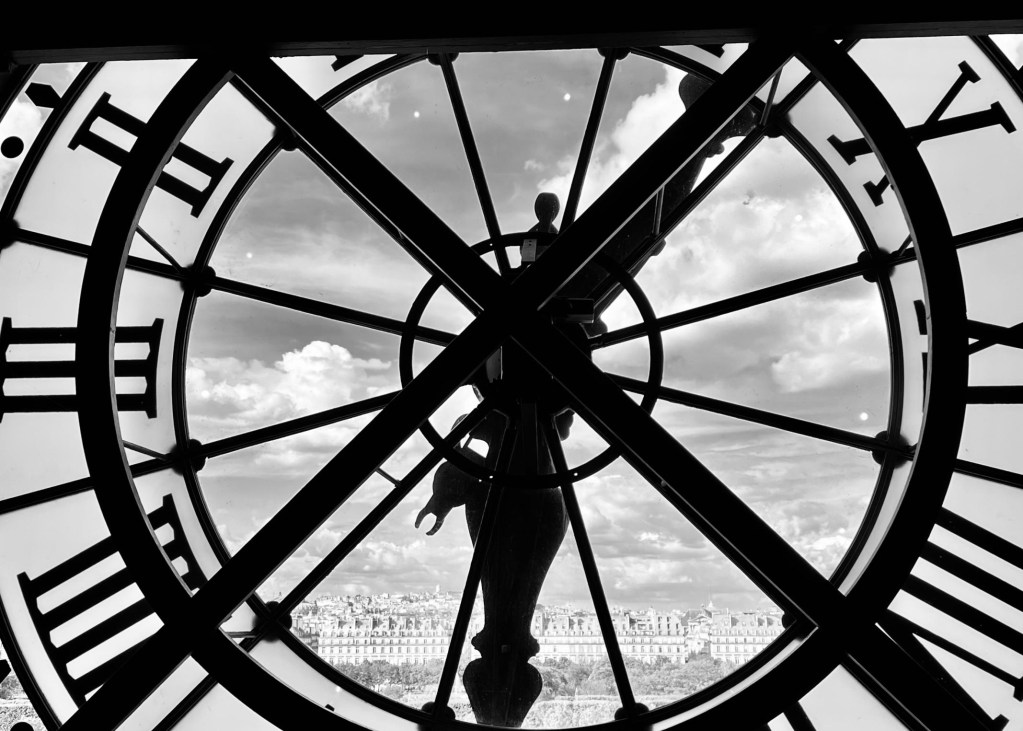 View through a large clock face, featuring a silhouette of a statue against a cloudy sky and a cityscape in the distance.