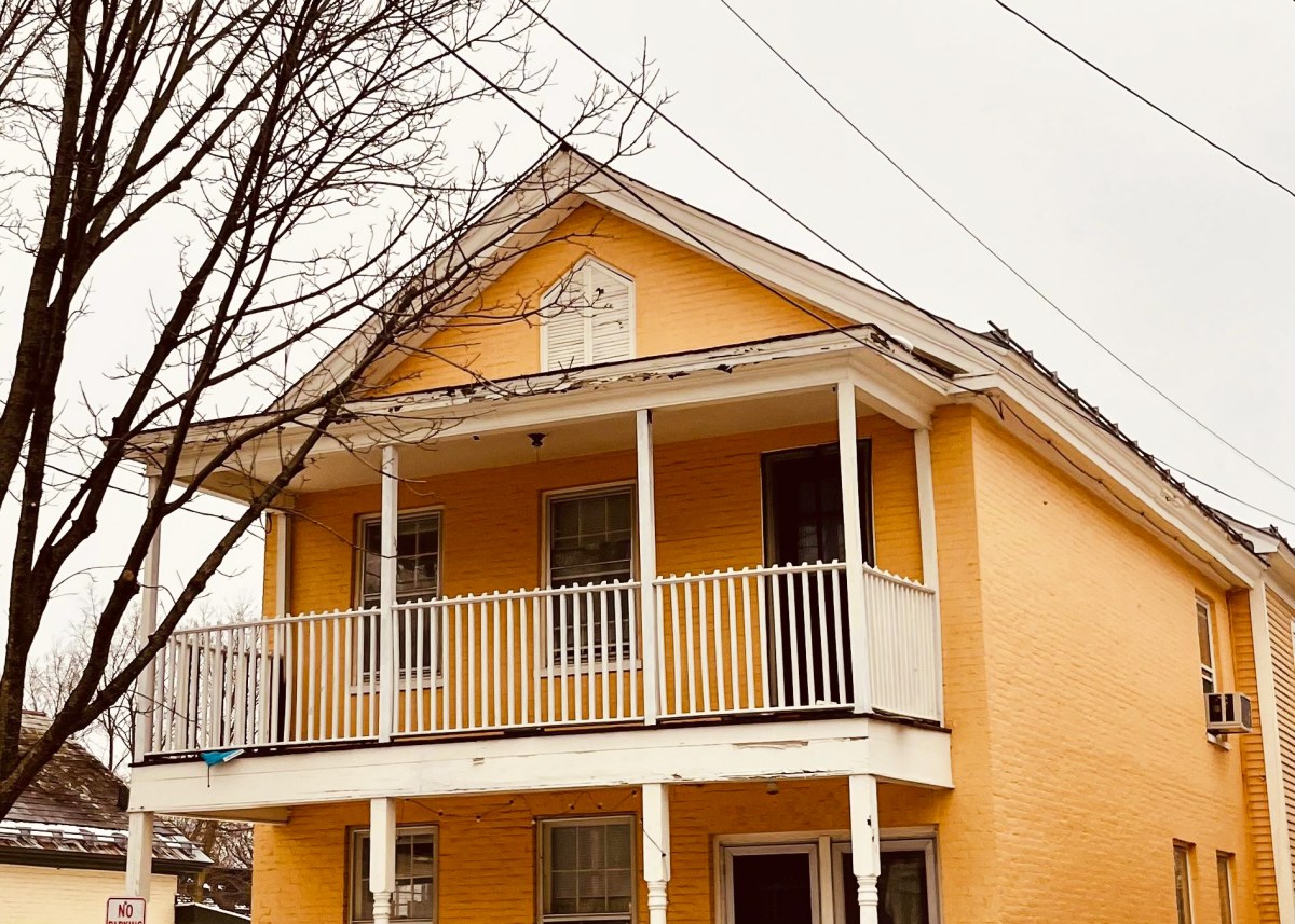 A two-story yellow house with white trim and a porch, featuring a front balcony and leafless trees nearby.