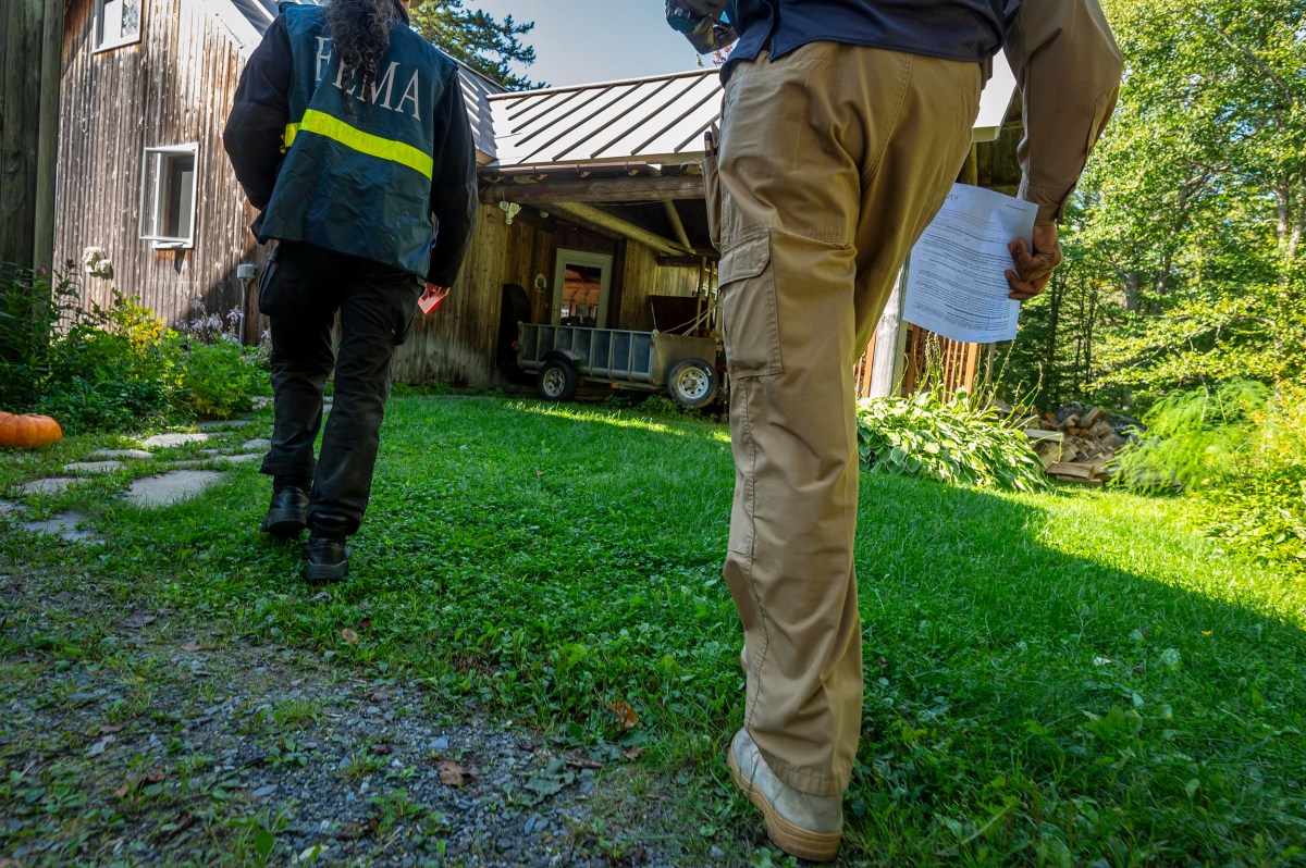 Two individuals, one wearing a FEMA jacket, walk beside a house with a shed in the background. One carries papers in their hand. Green grass and plants surround the area.