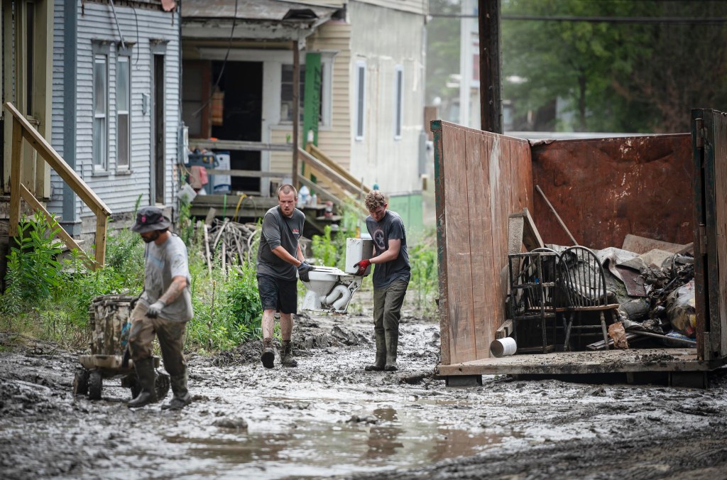 Three people working in a muddy, flooded alleyway clean up and remove debris into a large dumpster. One person pushes a wheelbarrow, while the other two carry items through the sludge.