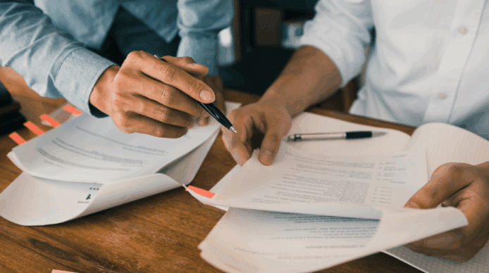 Two paper contracts open on a desk with the hands of two people visible