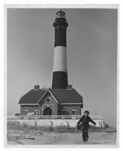 Black-and-white photograph of a lighthouse, patterned in bulky stripes. In the foreground, a young boy runs on the beach toward the camera.