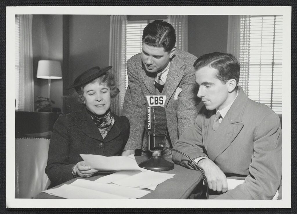 A black and white photograph shows Hallie Flanagan sits at a table with two men speaking into a microphone.