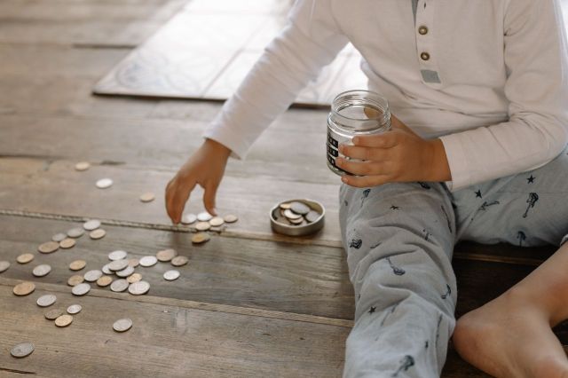 A child sitting on a wooden floor, holding a jar and picking up coins scattered around them.