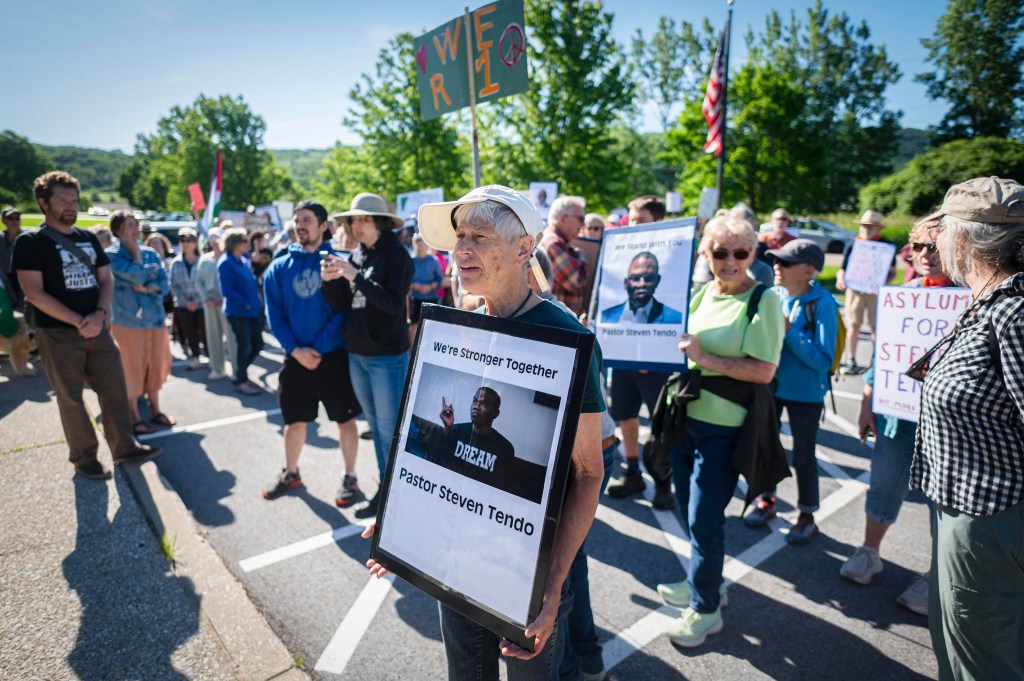 A group of people standing outdoors holding signs, including one featuring a photo of Pastor Steven Tendo and the text, "We're Stronger Together." Trees and blue sky are visible in the background.