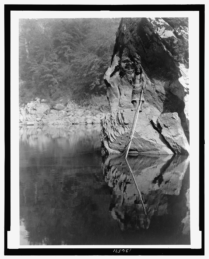 A black-and-white photograph depicting a Yurok man standing on a rock out in the water, fishing.
