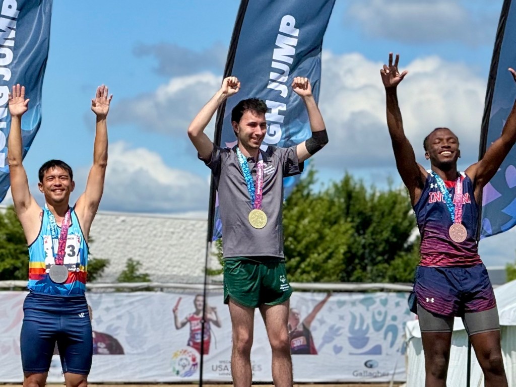 Three male athletes stand on a podium with medals, raising their arms in celebration after a long jump competition. Two event flags are visible in the background.