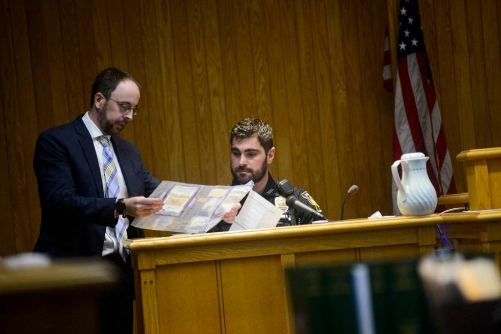 A man in a suit shows documents to a uniformed officer seated in a wooden witness stand in a courtroom with an American flag in the background.