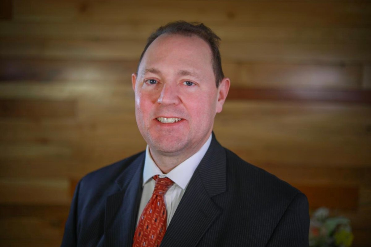 Man in a dark pinstripe suit with a red patterned tie stands in front of a wooden wall, smiling slightly at the camera.
